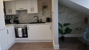Una cocina compacta con armarios blancos, un horno empotrado, un lavamanos y una cafetera. En el comedor contiguo, hay una planta en maceta y una mesa de vidrio.
