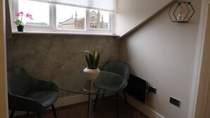 Un pequeño comedor con una mesa de vidrio y dos sillas, una planta en maceta, y vista a un edificio a través de una ventana.