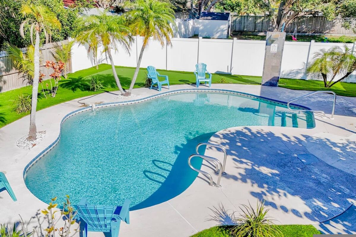 A rectangular swimming pool with a curved edge, surrounded by a white fence, and shaded by palm trees.