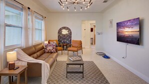 A modern living room with a brown leather sofa, a patterned rug, a chandelier, and a painting of a sunset on the wall.