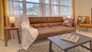 A living room with a brown leather sofa, a patterned throw blanket, a small wooden table with a lamp, and a patterned rug.