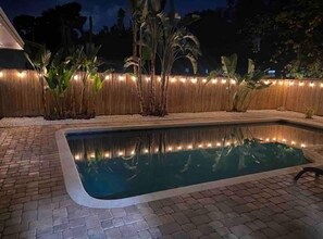 A swimming pool with a brick border, surrounded by plants and a bamboo fence.