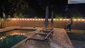 Zona de piscina con sillas de descanso y sombrillas, rodeada por una valla de madera y luces colgantes.