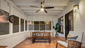 Une terrasse couverte avec une table et des chaises en bois, un ventilateur de plafond et de grandes fenêtres.