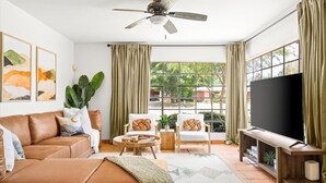 Un salon avec un canapé marron, deux fauteuils, une table basse, un téléviseur posé sur un meuble en bois et une grande fenêtre avec des rideaux.