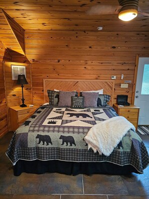 Un dormitorio de cabaña de madera con una cama, una mesita de noche, una lámpara, un termostato, un cartel de 'No Fumar' y una puerta con ventana.