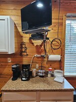 Una cocina con un televisor montado en la pared, una cafetera, una jarra de café, una taza, un portavasos para café y una cesta.