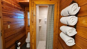 Une salle de bain rustique, aménagée dans une cabane en bois, avec des toilettes, une douche et des serviettes rangées sur un porte-serviettes.