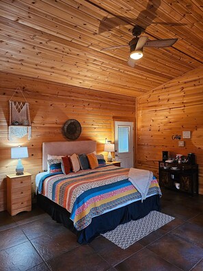 Ein gemütliches Schlafzimmer in einer Blockhütte mit Holzbalkendecke, einem Bett mit farbenfroher Steppdecke, einem Nachttisch mit Lampe und einer Kaffeestation.