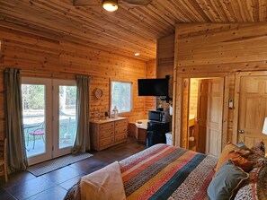 A wooden cabin room with a bed, a desk with a computer, a television, and a door leading outside.