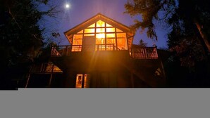 Una cabaña de madera con un ventanal grande, rodeada de árboles y una luna en el cielo.