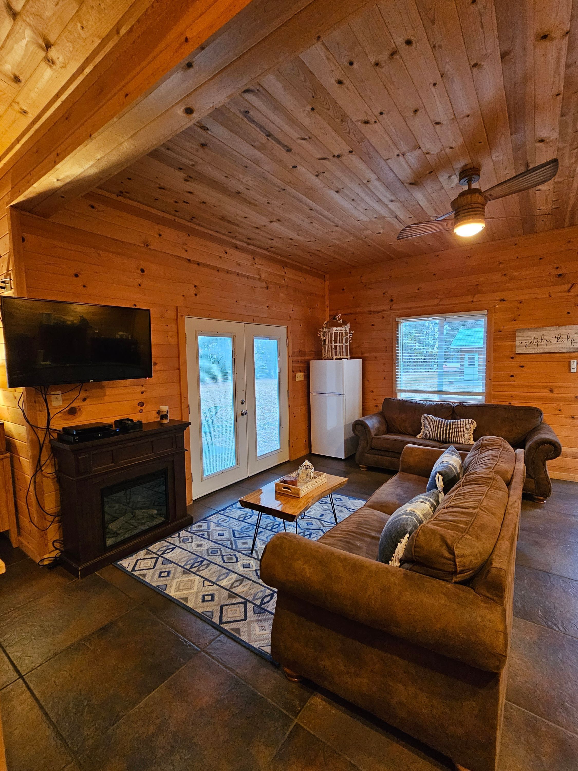 L’intérieur chaleureux d’un chalet avec une cheminée, un canapé, un téléviseur et un ventilateur de plafond.
