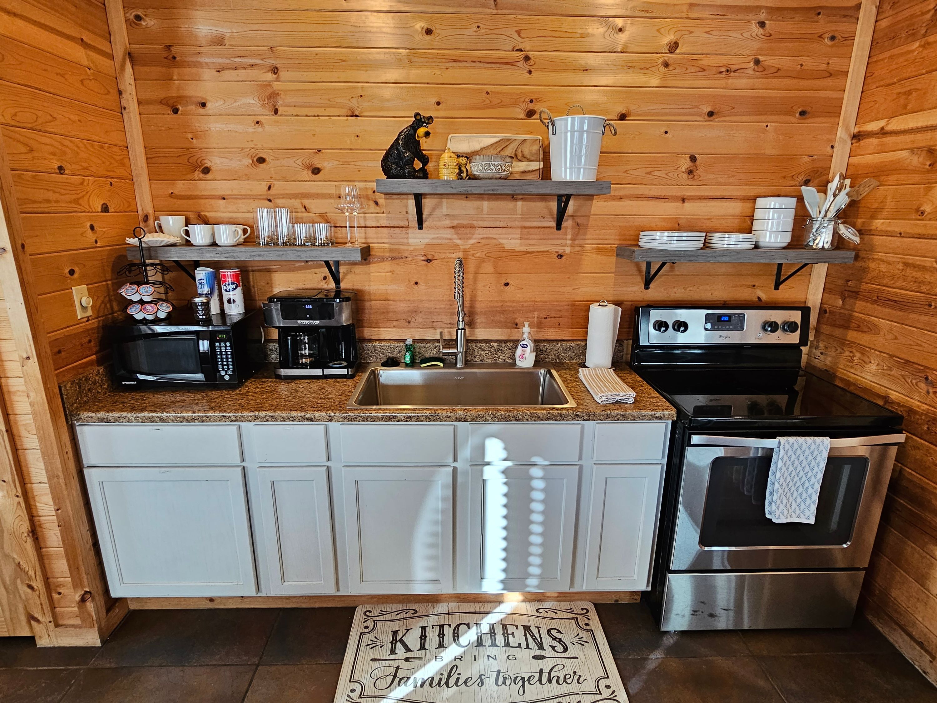 A compact kitchen with wooden walls, a stainless steel sink, a coffee maker, a stove, and a small shelf with kitchen utensils.