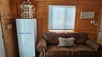 A wooden cabin interior with a white refrigerator, a brown sofa with a patterned pillow, and a window with blinds.