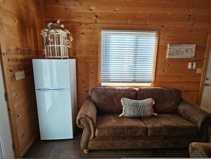 Interior de una cabaña de madera con un refrigerador blanco, un sofá marrón y un cojín estampado, además de una ventana con persianas.