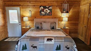 A wooden cabin bedroom with a bed, two bedside tables, a door, and a wall-mounted painting.