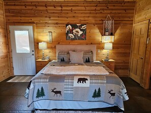 Un dormitorio de cabaña de madera con una cama, dos mesitas de noche, una puerta y un cuadro colgado en la pared.