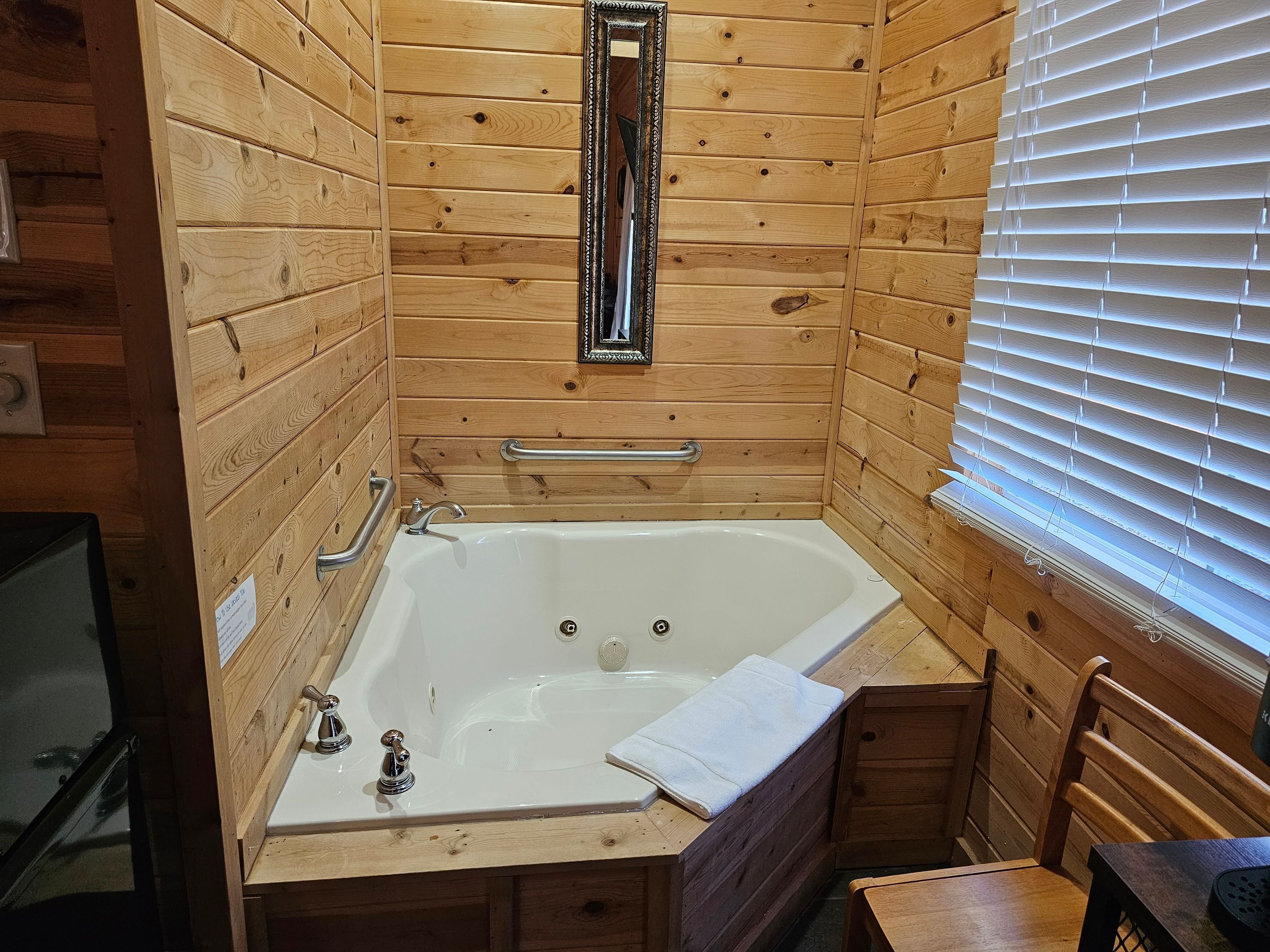 Ein Badezimmer einer Holzhütte mit einer Badewanne, Holzwänden, einem Spiegel und einem Fenster mit Jalousien.