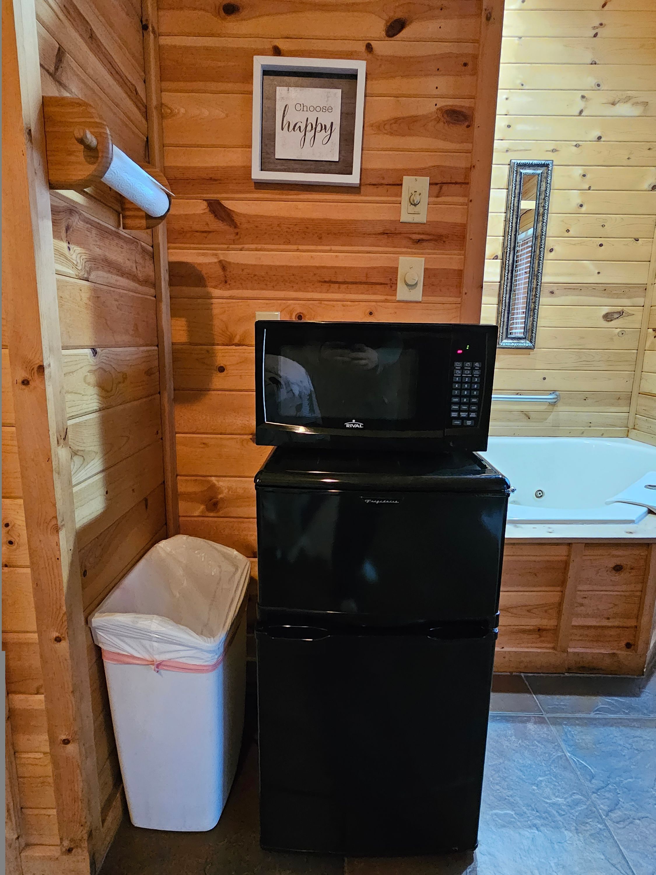 Habitación compacta con microondas sobre un refrigerador, un bote de basura, una cita motivacional enmarcada en la pared y una ventana con reja de metal.