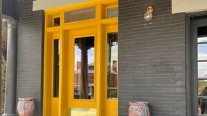 Entrada moderna de un edificio con una puerta amarilla, paredes de ladrillo negro y dos jarrones decorativos a cada lado.