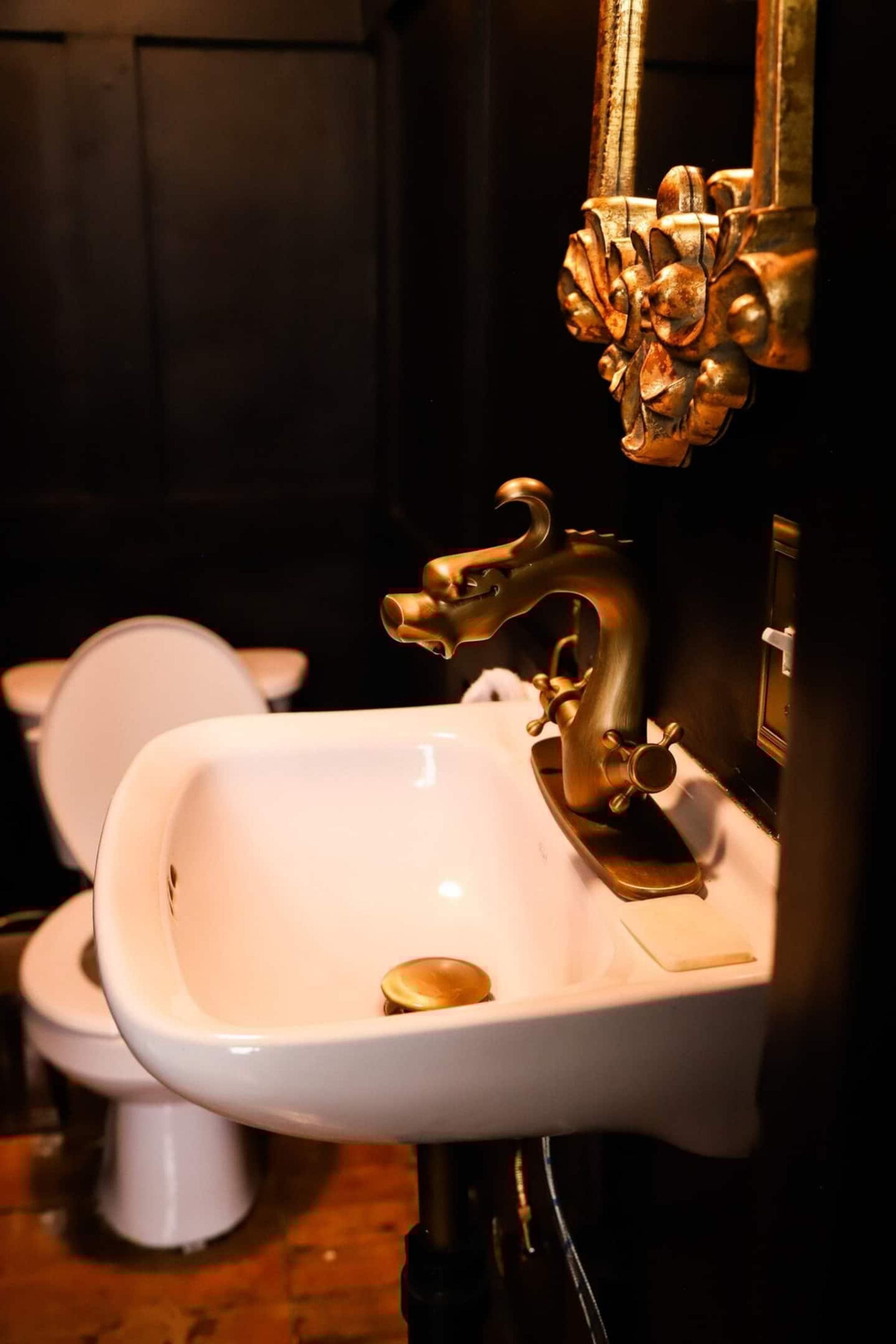 Ein Badezimmer mit einem weißen Waschbecken und einem goldenen Wasserhahn, einer weißen Toilette und einem Spiegel mit aufwendiger Rahmung.