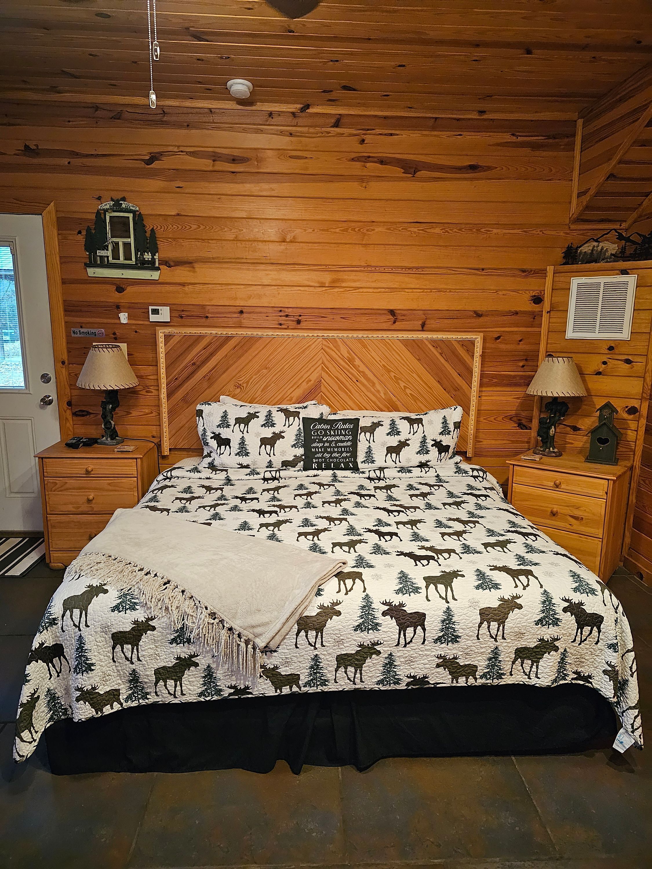 Ein Schlafzimmer einer Holzhütte mit einem Bett, das Elche und Waldmotive auf der Bettwäsche zeigt, zwei Nachttische mit Lampen, einem 'Nichtraucher'-Schild und einer kleinen Ablagefläche mit einem dekorativen Gegenstand.