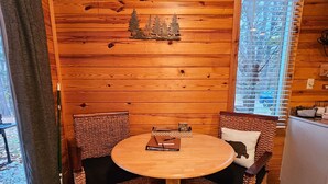 A wooden-paneled room with a round table and two wicker chairs, a window with blinds, and a small shelf with a decorative item. - Honey Bear Haven Suite 1 (Eureka Springs)