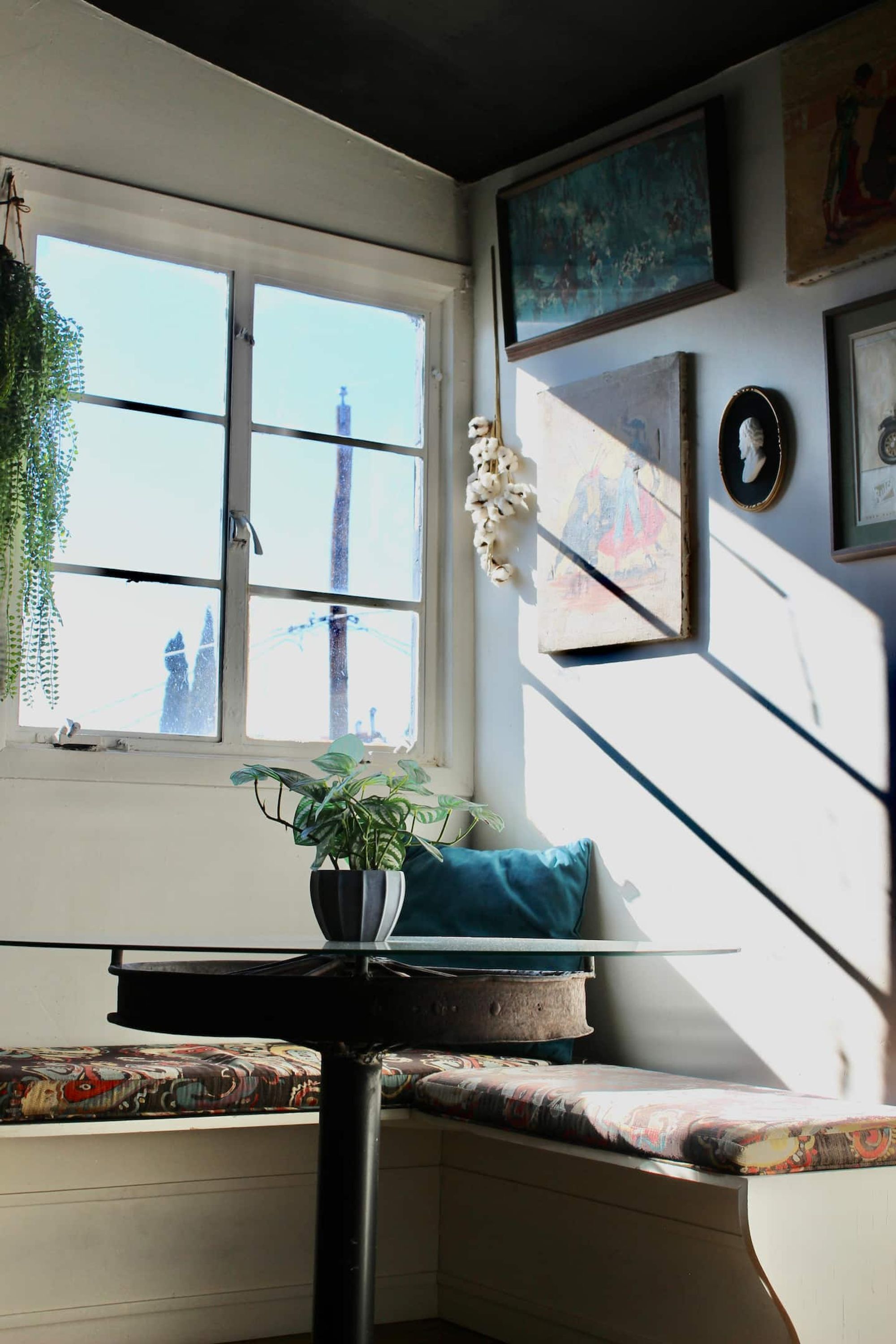 Een kamer met een raam, een tafel met een plant, een blauw kussen en kunstwerken aan de wanden.
