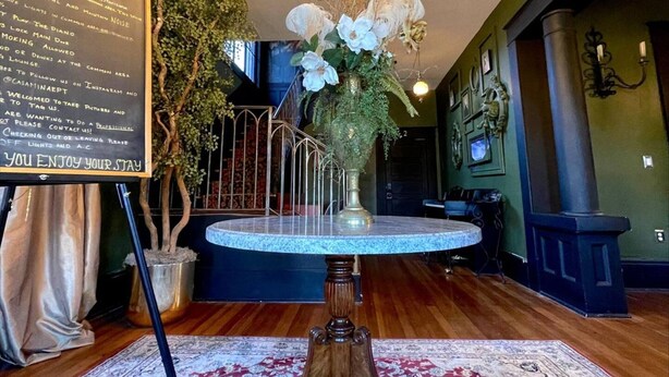 A lobby with a chandelier, a round table with a vase of flowers, a signboard with information, and a potted plant. - Casa Mina 5 Central Rio Grande Historic Dist (El Paso)
