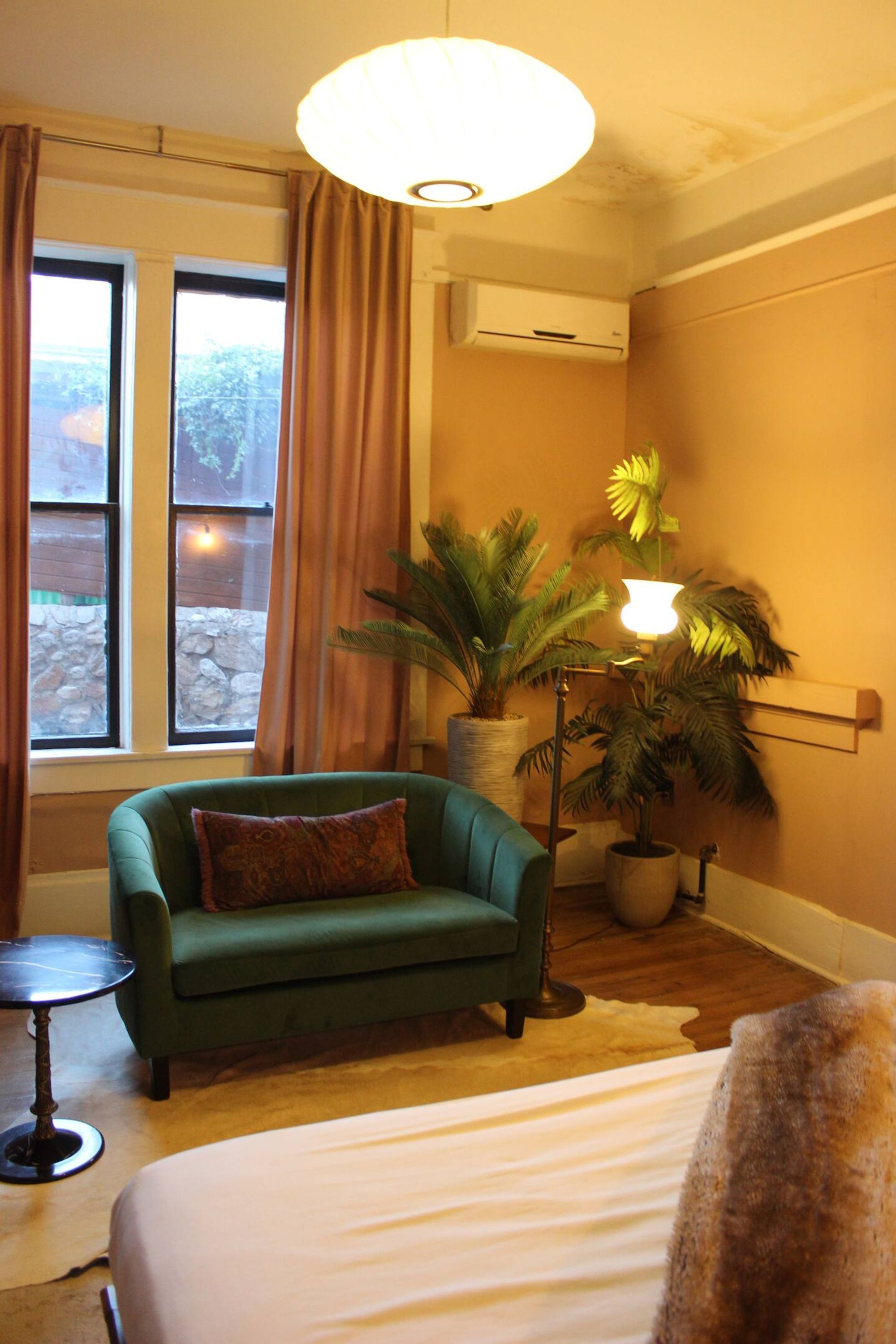 Une pièce comprenant un lit, un fauteuil vert, une petite table, une plante en pot, une lampe sur pied et une fenêtre avec des rideaux.