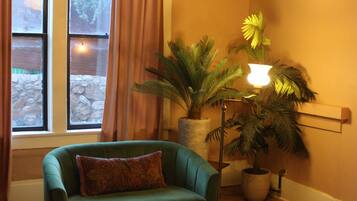 Une pièce comprenant un lit, un fauteuil vert, une petite table, une plante en pot, une lampe sur pied et une fenêtre avec des rideaux.