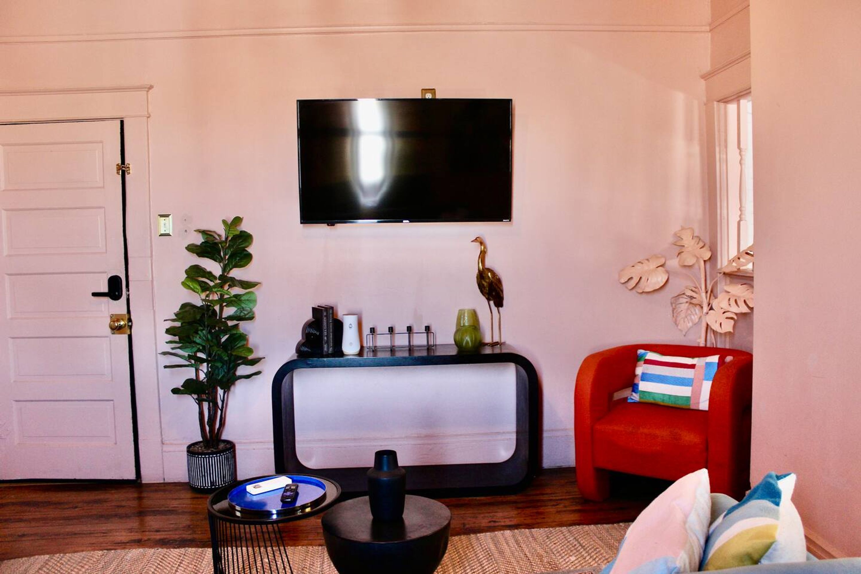 Uma sala de estar com televisão de tela plana na parede, uma poltrona vermelha com almofada listrada, um vaso de planta e uma pequena mesa lateral com um controle remoto.