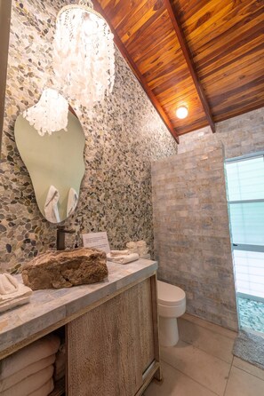 A bathroom with a stone mosaic wall, a wooden ceiling, a large mirror, and a rustic sink. - Casa Bonita - 5BR With Pool Walk to Beach (Nosara)