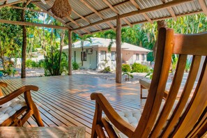 A wooden deck with chairs, a covered area with a corrugated metal roof, and a view of a building with a white roof and beige walls. - Casa Bonita - 5BR With Pool Walk to Beach (Nosara)
