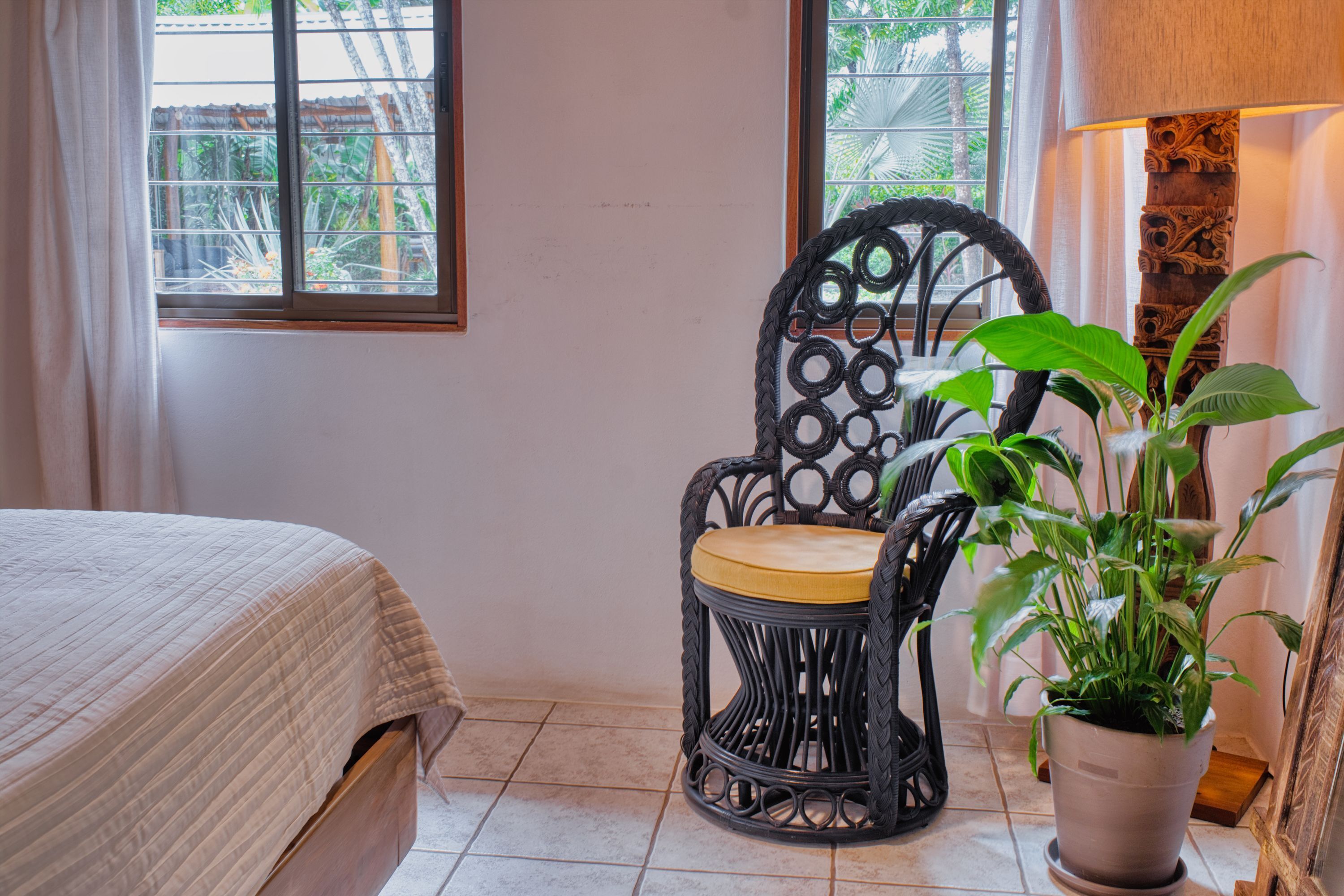 A bedroom with a bed, a wicker chair with a cushion, a potted plant, and a lamp.