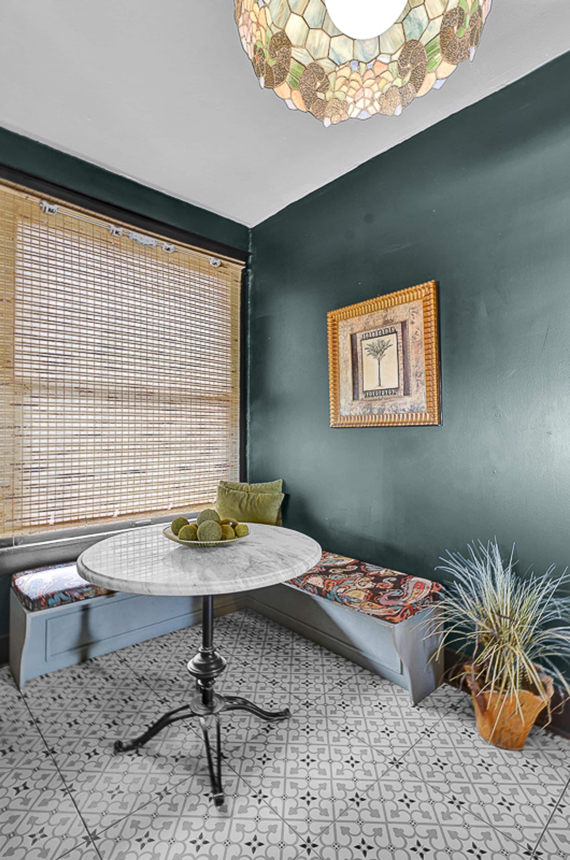 Um quarto com uma mesa redonda, um banco, um vaso de planta, um quadro pendurado na parede e uma janela com persianas.