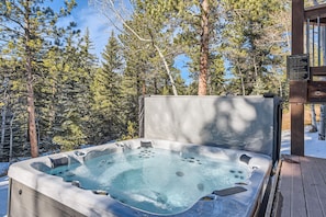 A hot tub in a snowy forest setting with a wooden deck and a safety sign. - Creekside Mountain Views Hot Tub (Evergreen)