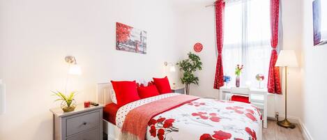 Un dormitorio con cama, mesitas de noche, una lámpara, una ventana con cortinas, una planta, un reloj y un cuadro en la pared.