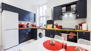 Une cuisine moderne avec des meubles de couleur sombre, un réfrigérateur blanc, un lave-linge et un coin repas avec une table blanche et des sets de table rouges.