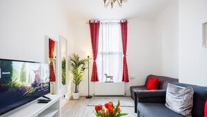 Un salon moderne avec un téléviseur à écran plat, un canapé gris, des coussins rouges, une table basse blanche ornée de tulipes et une grande fenêtre avec des rideaux rouges.