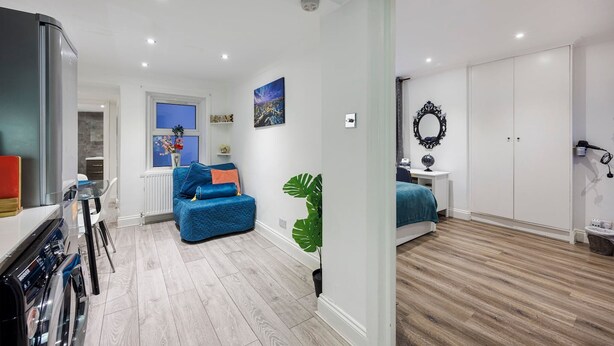 A modern apartment with a kitchen, living area, and bedroom. It features a blue sofa, a large plant, and a wardrobe. - Cute and Cosy Apartment in Kensington (London)