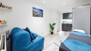 A modern living room with a blue sofa, a dining table with blue placemats, a cityscape painting, and a kitchen in the background. - Cute and Cosy Apartment in Kensington (London)