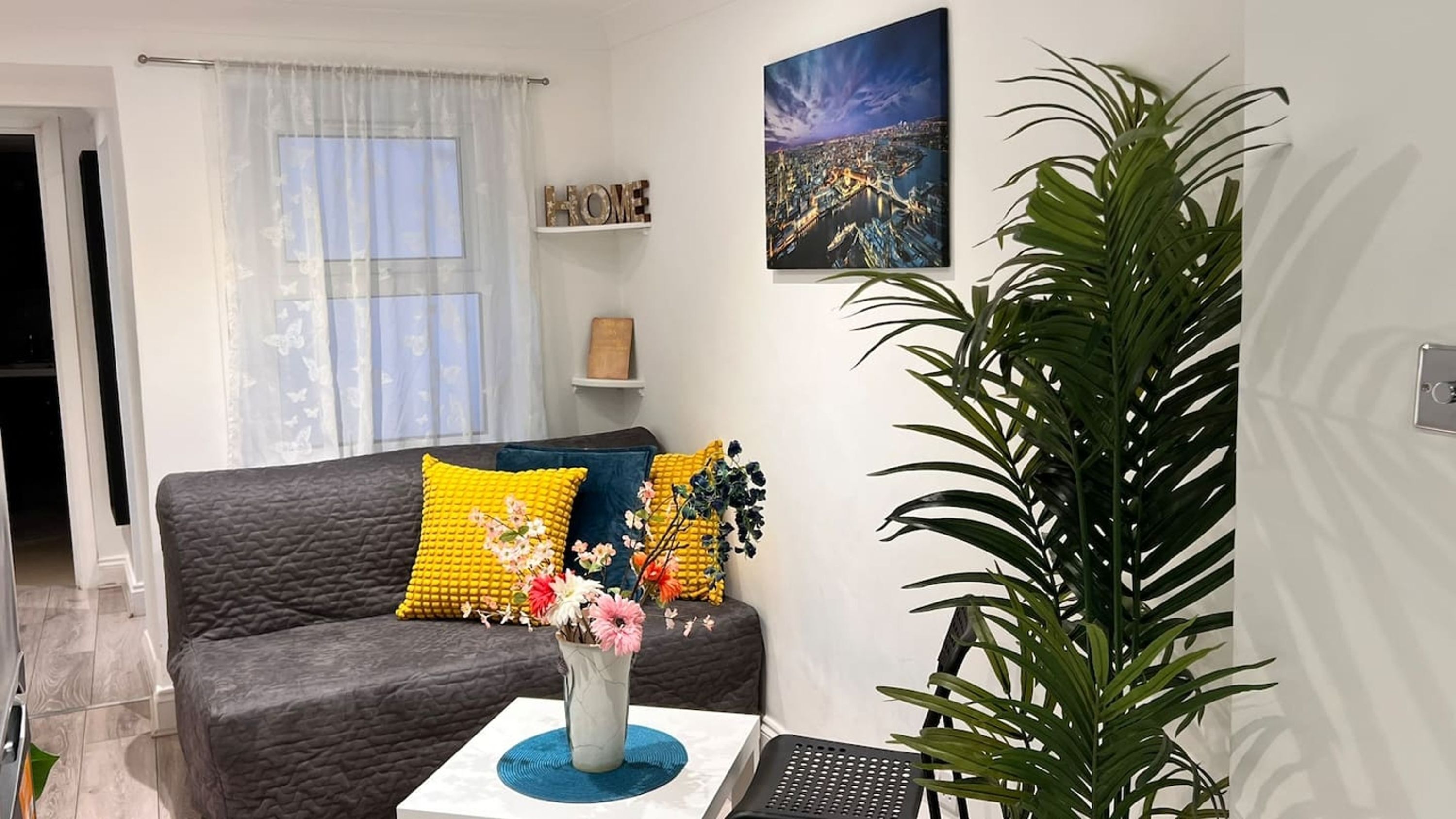 A modern living room with a grey sofa, yellow and blue cushions, a vase with flowers, a large potted plant, and a cityscape painting on the wall.