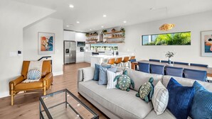 A modern living room with a sofa, armchair, and glass coffee table. A kitchen and dining area are visible in the background. - NEW 4 Bed 3 Bath Heated Salt Pool Walk2beach (Indian Rocks Beach)