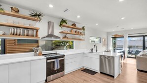 A modern kitchen with white cabinetry, wooden shelves, and a large window. - NEW 4 Bed 3 Bath Heated Salt Pool Walk2beach (Indian Rocks Beach)
