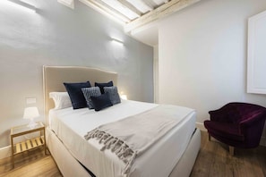 Una cama bien hecha con un cabecero, una mesita de noche con lámpara, y una silla morada en una habitación con suelo de madera y techo inclinado.
