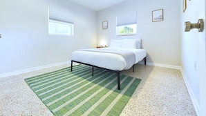 A bedroom with a bed, a ceiling fan, two windows with blinds, and a green striped rug.