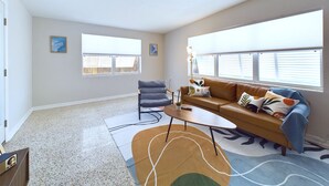 A spacious living room with a brown sofa, a gray armchair, and a coffee table. There are two windows with blinds, a ceiling fan, and a rug with abstract patterns.
