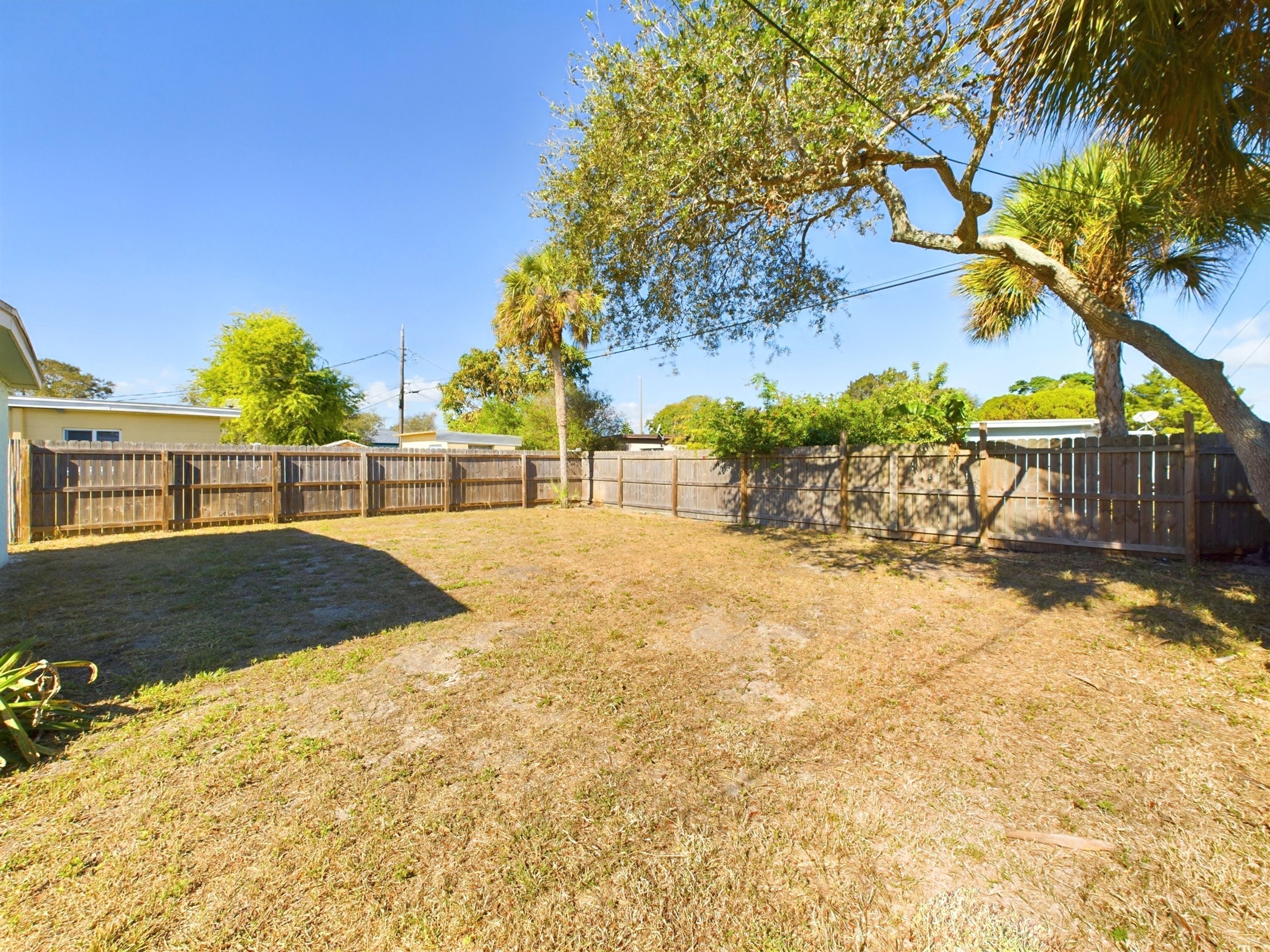 A fenced backyard with dry grass, a tree, and a house in the background.