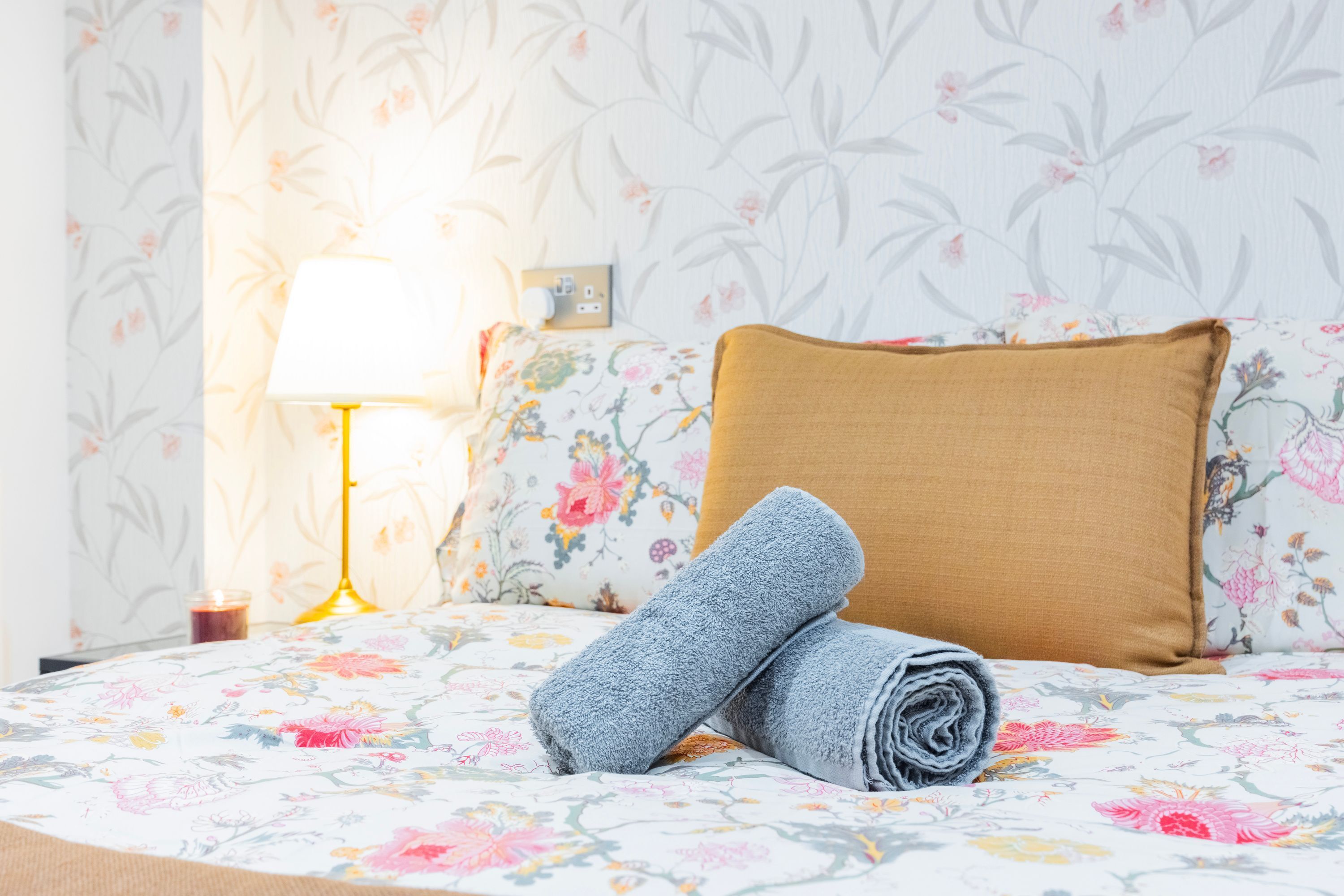 A neatly made bed with floral bedding, a rolled-up towel, a bedside lamp, and a wall with floral wallpaper.